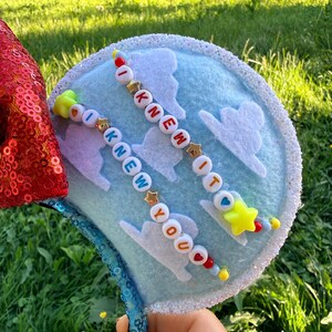 May include: A light blue headband ear featuring white felt clouds and two strands of colorful letter beads spelling out I KNEW IT and I KNEW YOU. The ear is bordered by white glitter trim and features a red sequined section on the side.