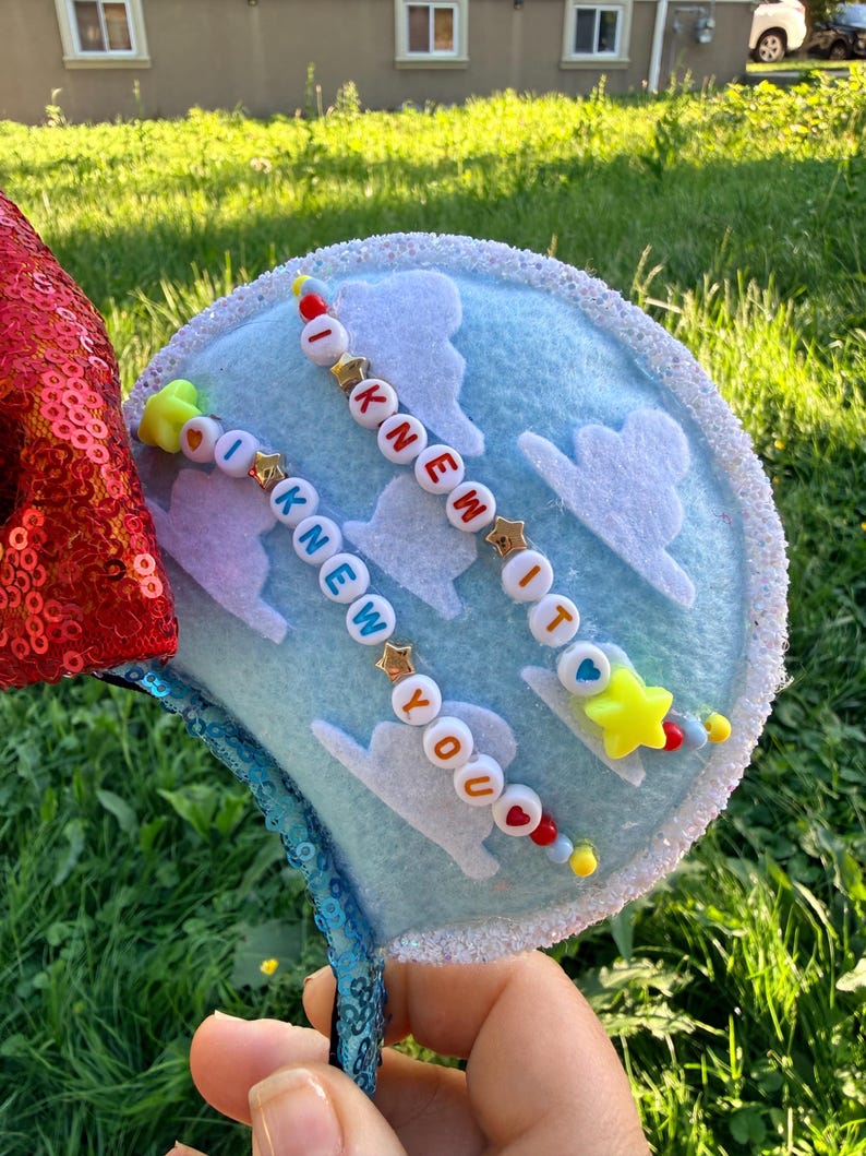 May include: A light blue headband ear featuring white felt clouds and two strands of colorful letter beads spelling out I KNEW IT and I KNEW YOU. The ear is bordered by white glitter trim and features a red sequined section on the side.