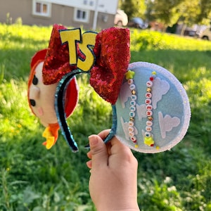 May include: A headband featuring two ears, one side decorated with a felt character face and the other with a light blue fabric featuring white cloud cutouts. A large red sequined bow sits in the center with yellow TS letters and beaded strands.