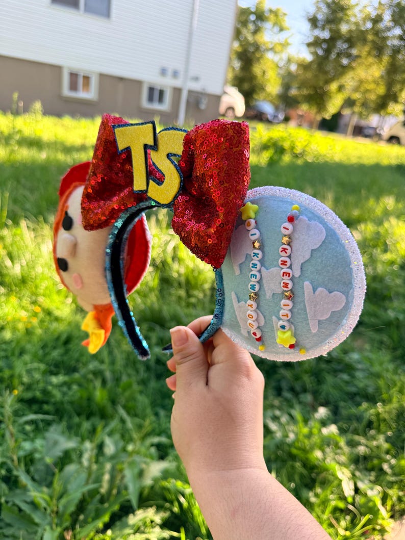 May include: A headband featuring two ears, one side decorated with a felt character face and the other with a light blue fabric featuring white cloud cutouts. A large red sequined bow sits in the center with yellow TS letters and beaded strands.