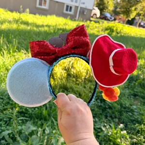May include: A headband featuring two circular ears, one light blue and one red with a white trim, topped with a red sequined bow. The headband band is covered in blue sequins. A small red hat is attached to the red ear, and yellow accents hang below.