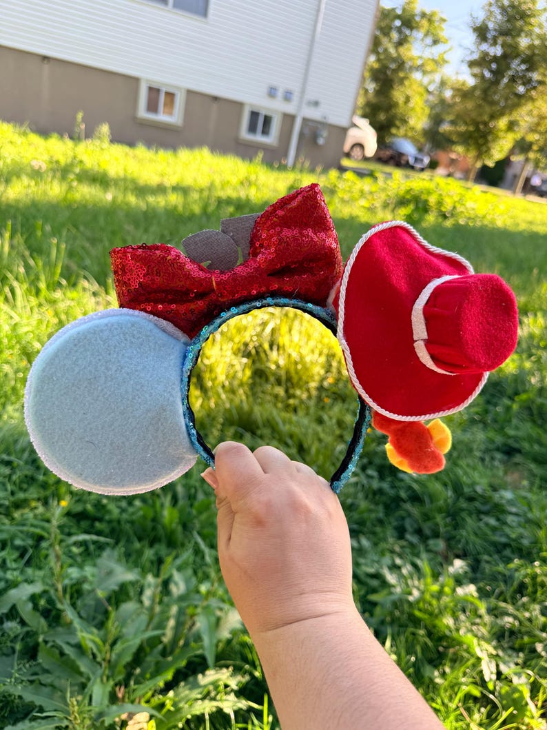 May include: A headband featuring two circular ears, one light blue and one red with a white trim, topped with a red sequined bow. The headband band is covered in blue sequins. A small red hat is attached to the red ear, and yellow accents hang below.