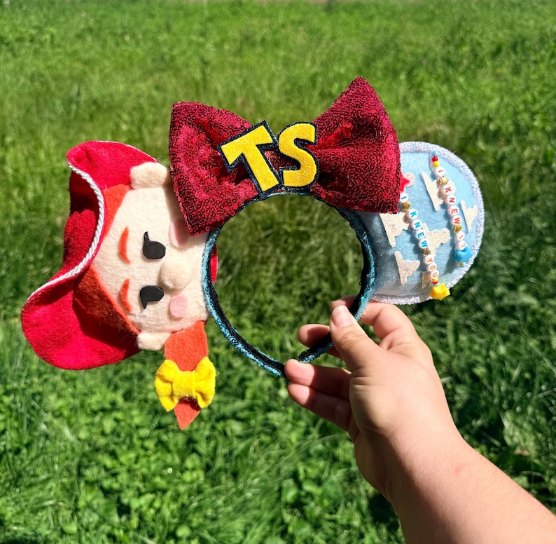 May include: A headband featuring a red sequined bow with yellow letters TS in the center. One side displays a felt character with red hair and a hat, while the other side is light blue with white cloud shapes and beaded strings reading I KNEW YOU.