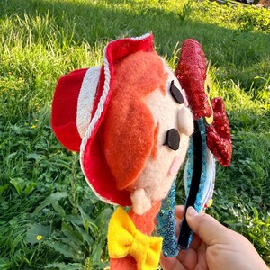 May include: A headband featuring a felt character head with bright orange hair, a red cowboy hat with white trim, and a yellow bow. The headband is adorned with a large, shiny red sequined bow on the side and light blue sequined fabric along the band.