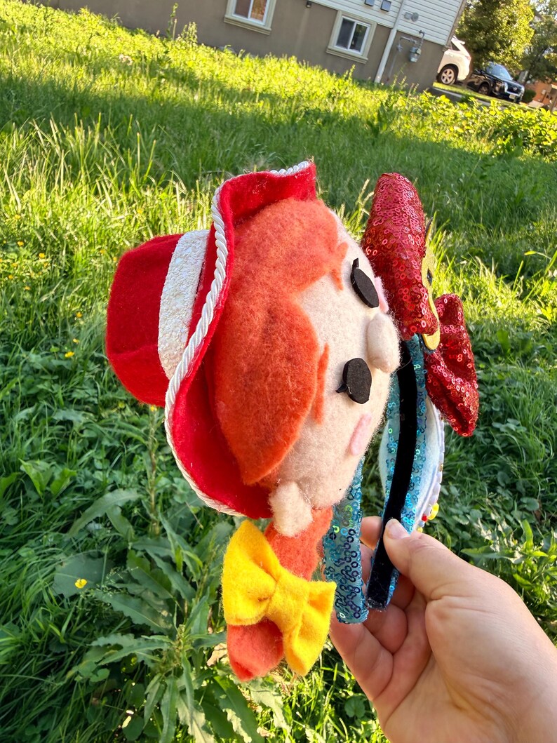 May include: A headband featuring a felt character head with bright orange hair, a red cowboy hat with white trim, and a yellow bow. The headband is adorned with a large, shiny red sequined bow on the side and light blue sequined fabric along the band.