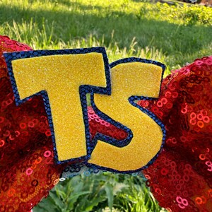 May include: A pair of headband ears covered in bright red sequins featuring large, yellow glittery letters T and S centered on the front. Each letter is outlined with a dark blue, textured border, creating a bold contrast against the red background.