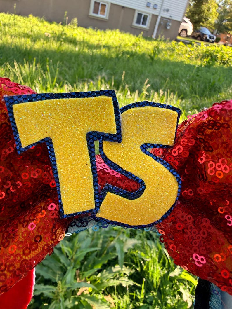 May include: A pair of headband ears covered in bright red sequins featuring large, yellow glittery letters T and S centered on the front. Each letter is outlined with a dark blue, textured border, creating a bold contrast against the red background.