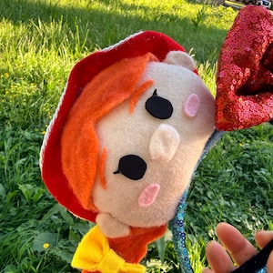 May include: A headband featuring a plush character head with orange hair, a red hat with white trim, and a yellow bow. One side has a red sequined ear, while the other side features a blue sequined band. The character has black eyes and pink cheeks.