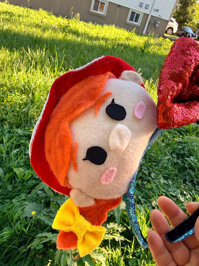 May include: A headband featuring a plush character head with orange hair, a red hat with white trim, and a yellow bow. One side has a red sequined ear, while the other side features a blue sequined band. The character has black eyes and pink cheeks.