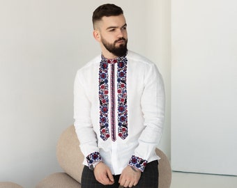 Peut inclure: Un homme portant une chemise en lin blanche avec des motifs floraux brodés en rouge, bleu et blanc. Il porte un pantalon noir à carreaux et des chaussures noires. Il se tient devant un mur blanc avec un coussin cylindrique beige derrière lui.