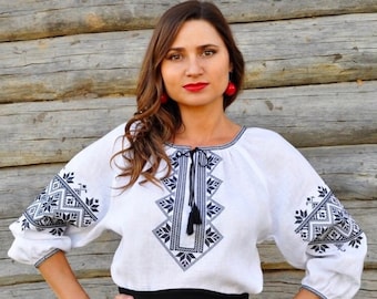 Puede incluir: Una blusa de lino blanca con patrones geométricos bordados en negro. La blusa tiene un escote redondo y mangas largas. La modelo lleva una falda negra.