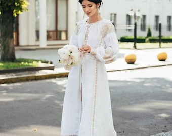 Könnte beinhalten: Eine Frau in einem weißen Leinenkleid mit goldener Stickerei und einem weißen Blumenstrauß. Das Kleid hat einen langen, fließenden Rock und ein tailliertes Oberteil. Die Frau steht auf einer Kopfsteinpflasterstraße vor einem weißen Gebäude.