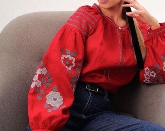 Könnte beinhalten: Eine rote bestickte Bluse mit langen Ärmeln und einem Blumendesign. Die Bluse hat einen runden Ausschnitt und einen Knopfverschluss. Das Model trägt eine blaue Jeans.