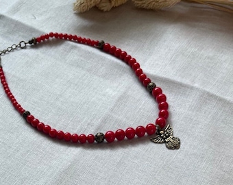 May include: A red beaded necklace with a silver angel pendant. The necklace is made of small, round beads and has a simple, elegant design.