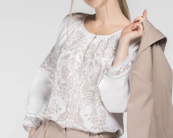 May include: A white blouse with beige embroidery featuring a floral and bird design. The blouse has long sleeves and is paired with beige pants. A beige jacket is held over the shoulder. The outfit is suitable for a casual or semi-formal occasion.