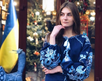Op de afbeelding: Een vrouw draagt een blauwe geborduurde blouse met witte bloemenpatronen en kwastjes. Ze staat voor een winkel met een bord waarop "Open Ukraine Now" staat in gele letters op een blauwe achtergrond.