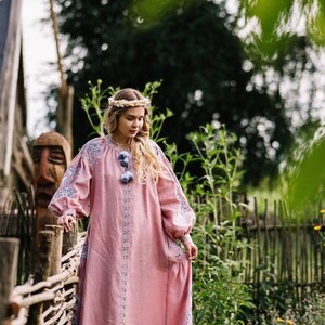 K&ouml;nnte beinhalten: Eine Frau in einem rosa bestickten Leinenkleid mit Blumenmuster und einer Blumenkrone steht vor einem geflochtenen Zaun.