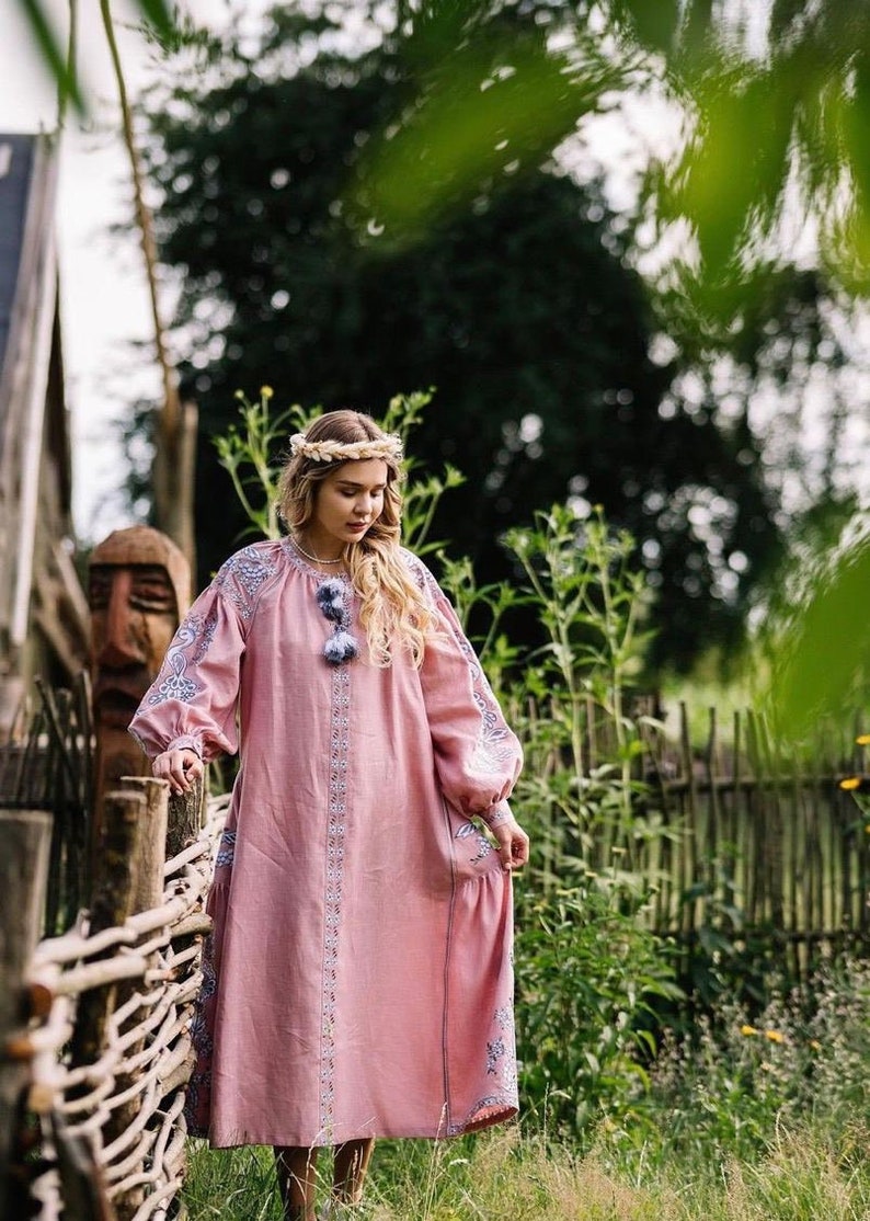 K&ouml;nnte beinhalten: Eine Frau in einem rosa bestickten Leinenkleid mit Blumenmuster und einer Blumenkrone steht vor einem geflochtenen Zaun.