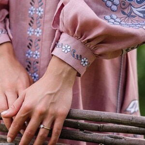 K&ouml;nnte beinhalten: Eine Frauenhand mit einem Ring mit einem klaren Stein, der auf einem Stapel Holzst&auml;mmen ruht. Die Hand tr&auml;gt ein rosa Leinenkleid mit gestickten Blumenmotiven.