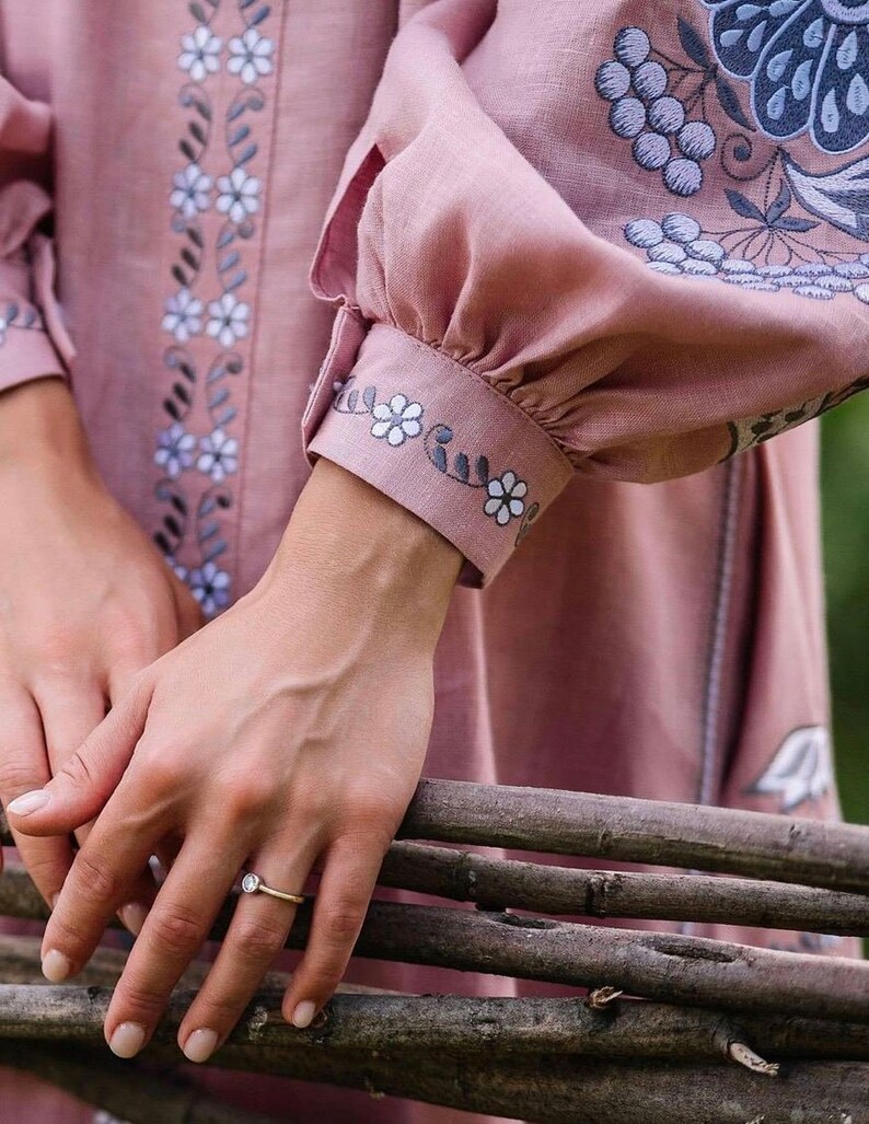 K&ouml;nnte beinhalten: Eine Frauenhand mit einem Ring mit einem klaren Stein, der auf einem Stapel Holzst&auml;mmen ruht. Die Hand tr&auml;gt ein rosa Leinenkleid mit gestickten Blumenmotiven.