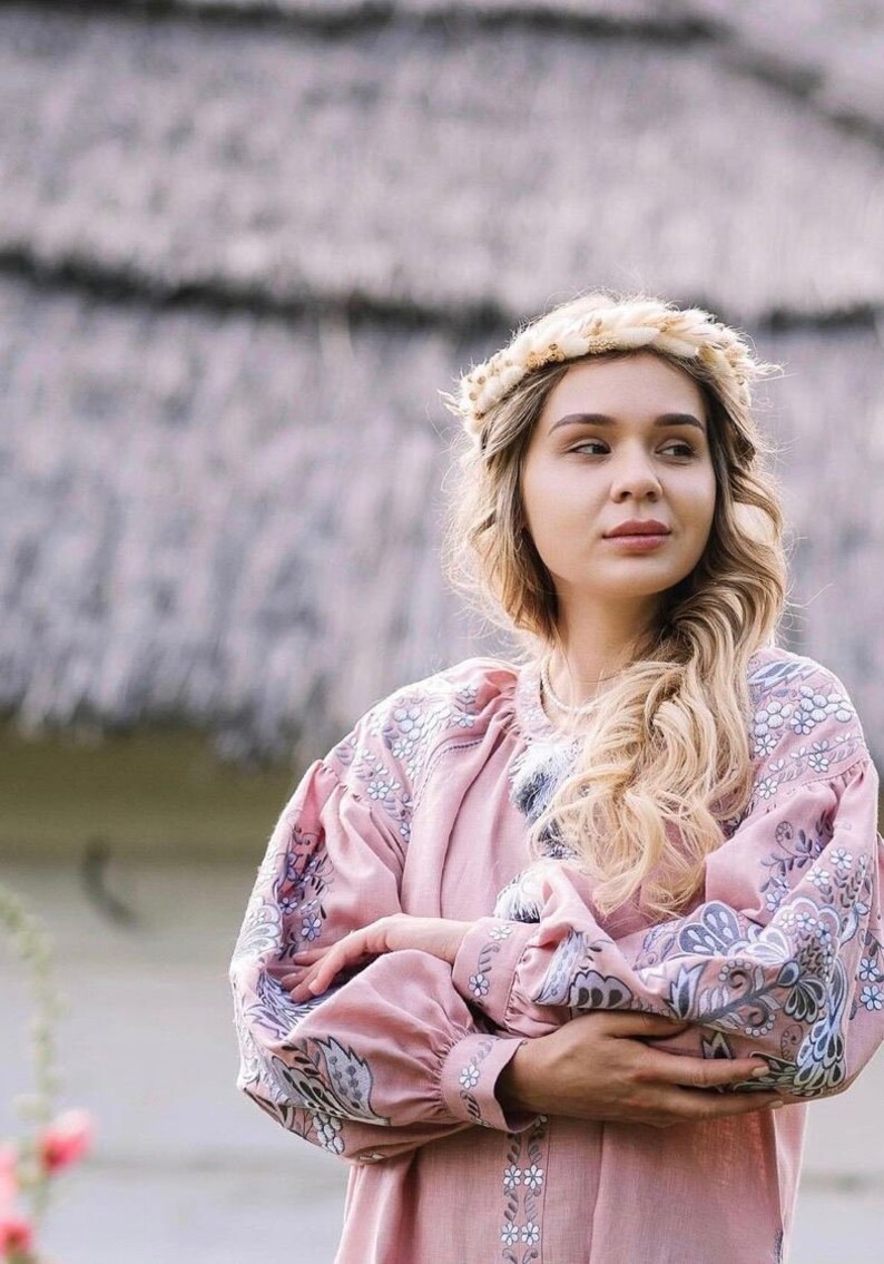 K&ouml;nnte beinhalten: Eine Frau tr&auml;gt eine rosa bestickte Bluse mit einem Blumenmuster. Sie tr&auml;gt ein gewebtes Stirnband und hat lange blonde Haare.