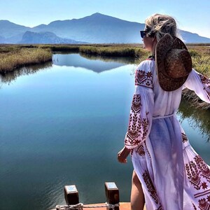 A woman wearing a white dress with red embroidery and a brown straw hat stands on a wooden boat. She is looking out at a body of water with mountains in the distance.