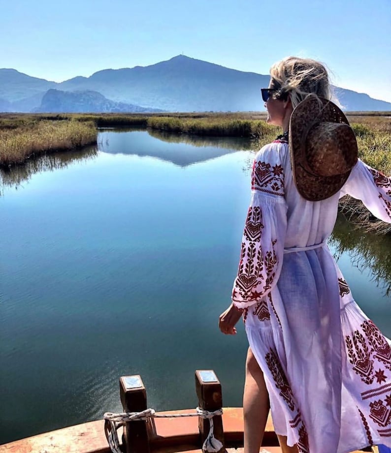 A woman wearing a white dress with red embroidery and a brown straw hat stands on a wooden boat. She is looking out at a body of water with mountains in the distance.