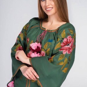 Puede incluir: Una blusa de lino verde con detalles florales bordados y borlas rosas y burdeos. La blusa tiene un cuello redondo y mangas largas.