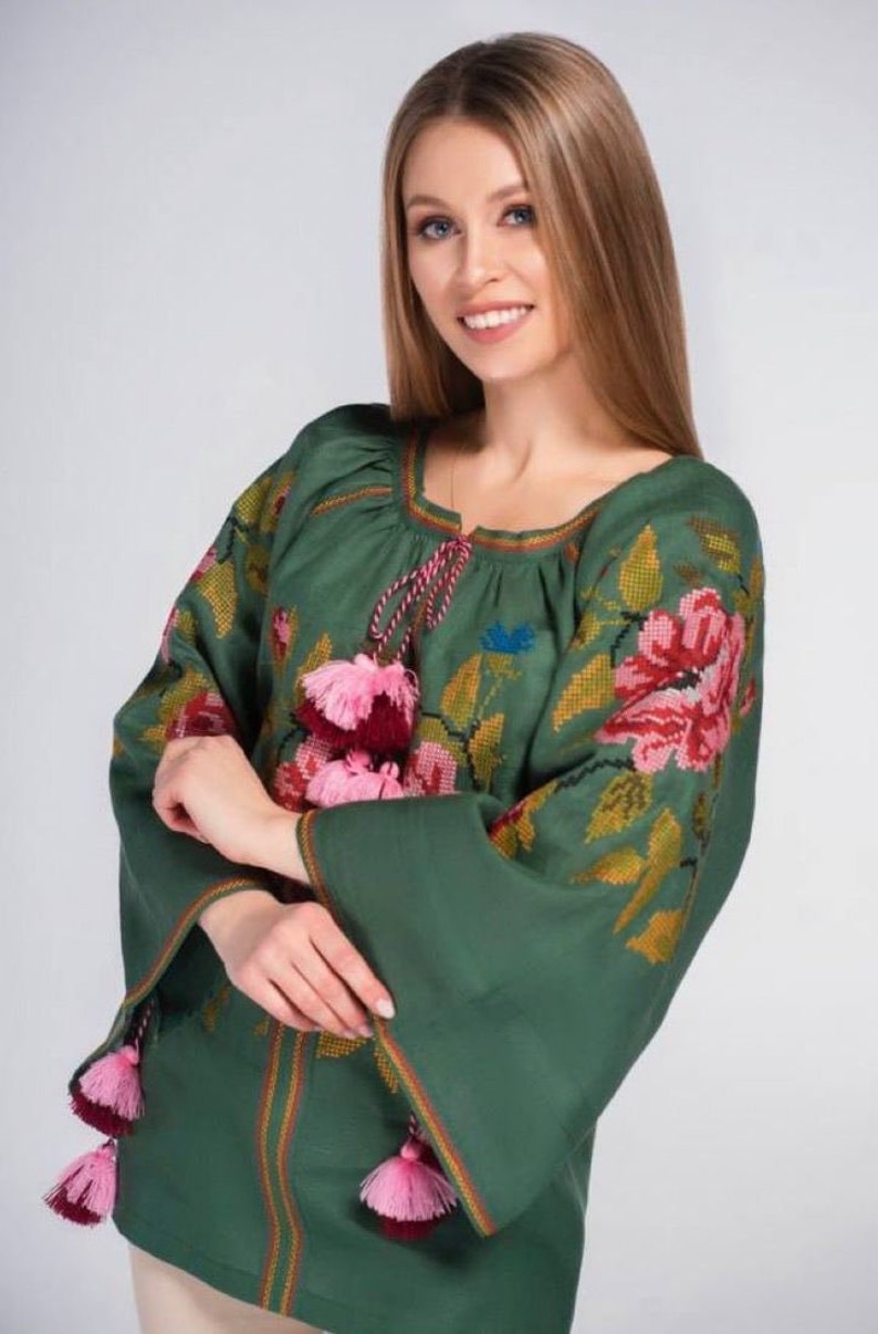 Puede incluir: Una blusa de lino verde con detalles florales bordados y borlas rosas y burdeos. La blusa tiene un cuello redondo y mangas largas.