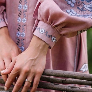 K&ouml;nnte beinhalten: Eine Frauenhand mit einem Ring mit einem klaren Stein, der auf einem Stapel Holzst&auml;mmen ruht. Die Hand tr&auml;gt ein lang&auml;rmeliges, rosafarbenes Leinenkleid mit gestickten Blumenmotiven.