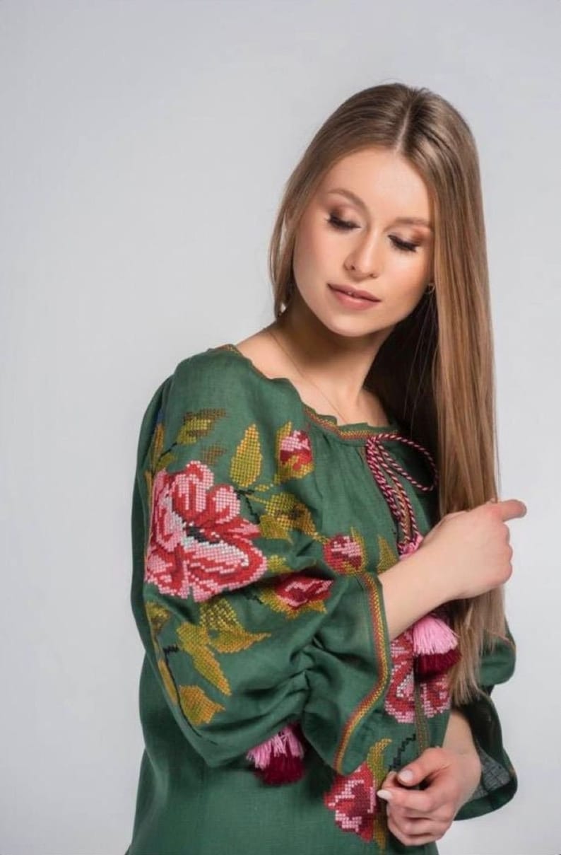 Puede incluir: Una blusa de lino verde con detalles florales bordados en rosa, rojo y amarillo. La blusa tiene mangas largas con borlas rosas y burdeos.