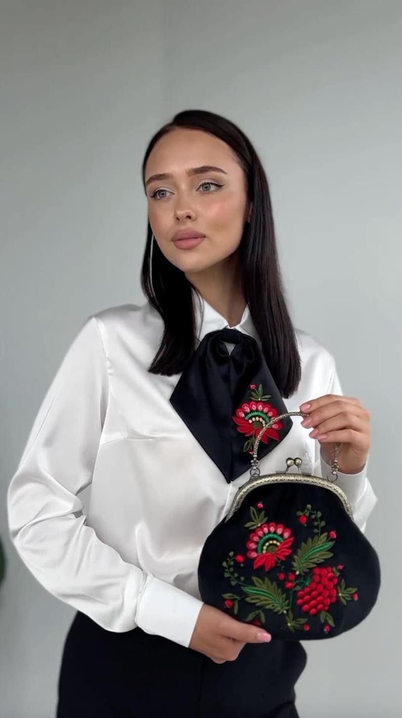 Pode incluir: Uma mulher vestindo uma blusa de cetim branco com uma gravata de la&ccedil;o de cetim preto e cal&ccedil;as pretas. Ela est&aacute; segurando uma bolsa de embreagem de veludo preto com um design de bordado floral em vermelho, verde e dourado.