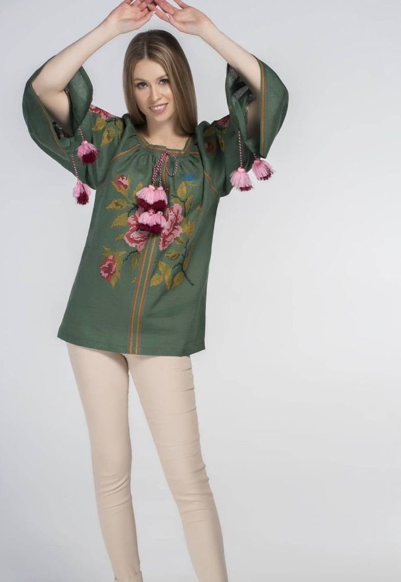 Puede incluir: Una blusa de lino verde con detalles florales bordados y borlas rosas y burdeos. La blusa tiene un escote redondo y mangas largas.