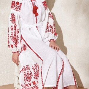 white linen dress with red embroidery. The dress has a round neckline, long sleeves, and a belt. The embroidery is in a floral pattern.