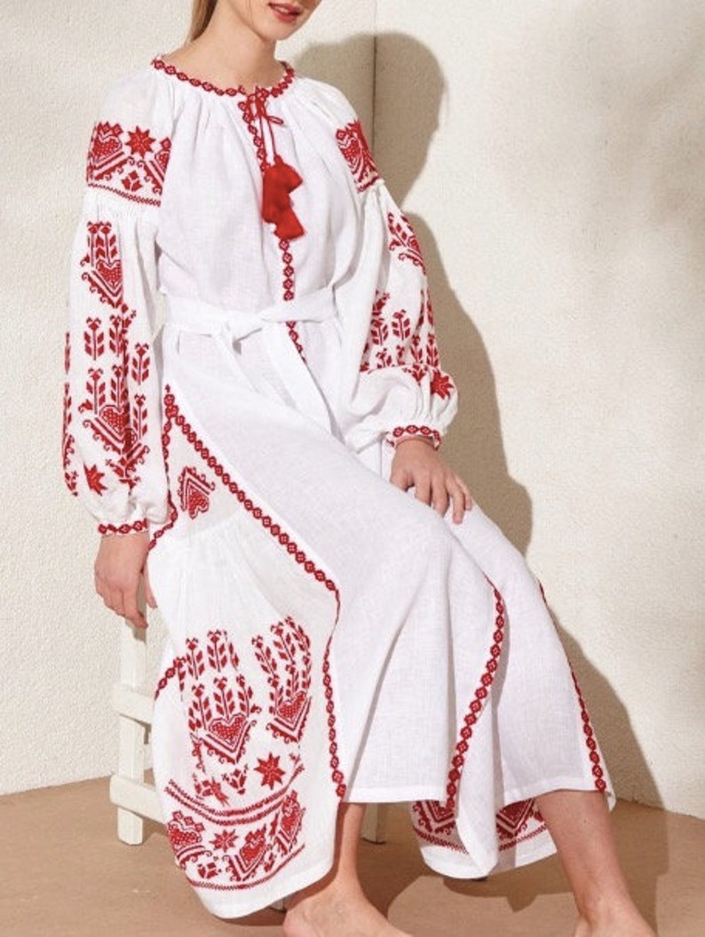white linen dress with red embroidery. The dress has a round neckline, long sleeves, and a belt. The embroidery is in a floral pattern.