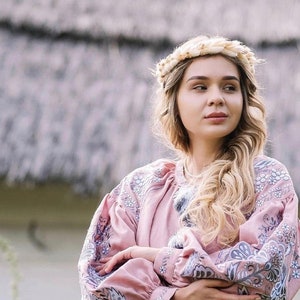 K&ouml;nnte beinhalten: Eine Frau mit langen blonden Haaren tr&auml;gt eine rosa bestickte Bluse mit Blumenmuster. Sie tr&auml;gt einen Kranz aus getrockneten Blumen im Haar.