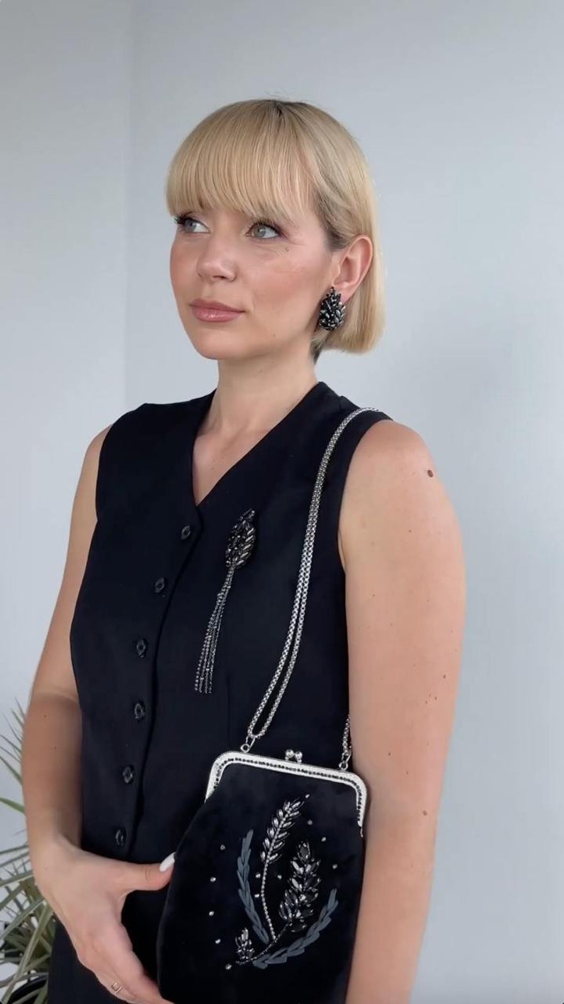 May include: A woman wearing a black sleeveless dress with a silver chain and a black beaded brooch. She is holding a black velvet clutch with a silver clasp and a beaded floral design.