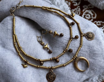 May include: A gold-toned necklace and earring set. The necklace features two strands with small bells, bird charms, and a crescent moon pendant. The earrings have bird charms and small bells. The jewelry is displayed on a gray cloth.