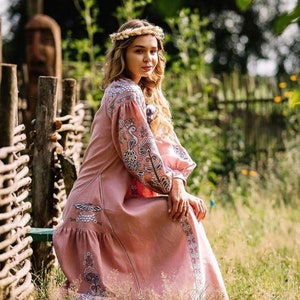 K&ouml;nnte beinhalten: Eine Frau in einem rosa bestickten Kleid mit langen &Auml;rmeln und einem Blumenmuster. Sie sitzt auf einer Holzbank in einem Garten.
