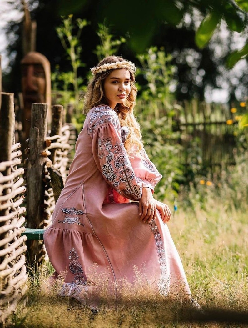 K&ouml;nnte beinhalten: Eine Frau in einem rosa bestickten Kleid mit langen &Auml;rmeln und einem Blumenmuster. Sie sitzt auf einer Holzbank in einem Garten.