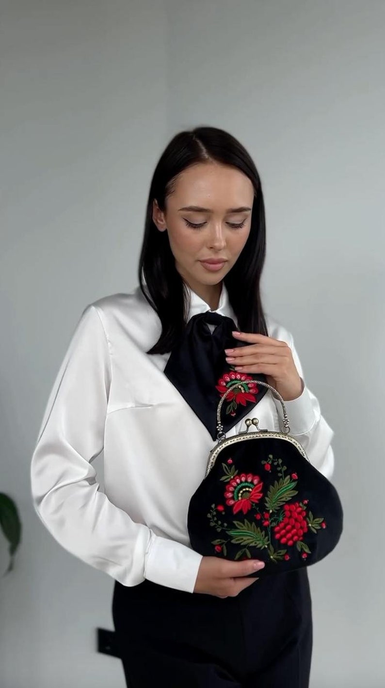 Pode incluir: Uma mulher vestindo uma blusa branca com uma gravata borboleta preta segura uma embreagem de veludo preto com flores bordadas vermelhas e verdes.