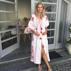May include: A woman wearing a white linen dress with red embroidery and tassels. The dress has a long, flowing skirt and a fitted bodice. She is standing in front of a door with a gray frame.