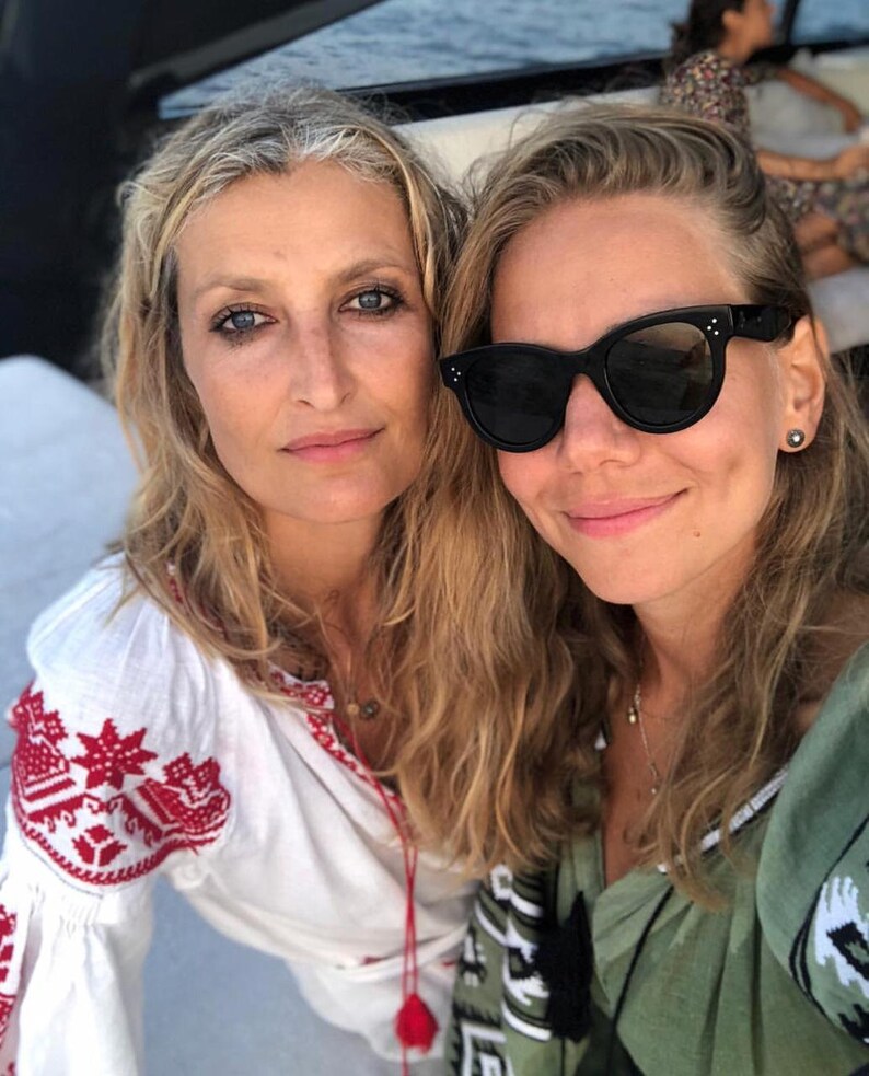 May include: Two women are smiling for a selfie. The woman on the left is wearing a white embroidered blouse with red accents. The woman on the right is wearing a green shirt with black and white embroidery and black sunglasses.