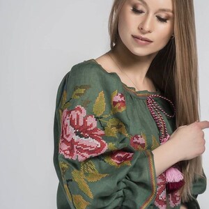 Puede incluir: Una blusa de lino verde con flores bordadas de color rosa y rojo. La blusa tiene un escote fruncido y mangas largas con borlas.