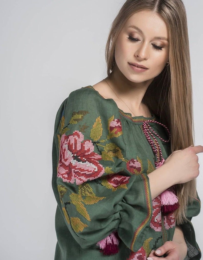 Puede incluir: Una blusa de lino verde con flores bordadas de color rosa y rojo. La blusa tiene un escote fruncido y mangas largas con borlas.