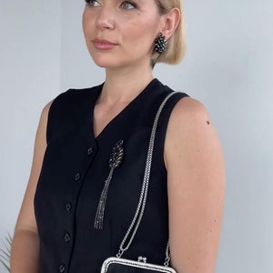 May include: A woman wearing a black sleeveless dress with a silver chain and a black beaded brooch. She is holding a black velvet clutch with a silver clasp and a beaded floral design.