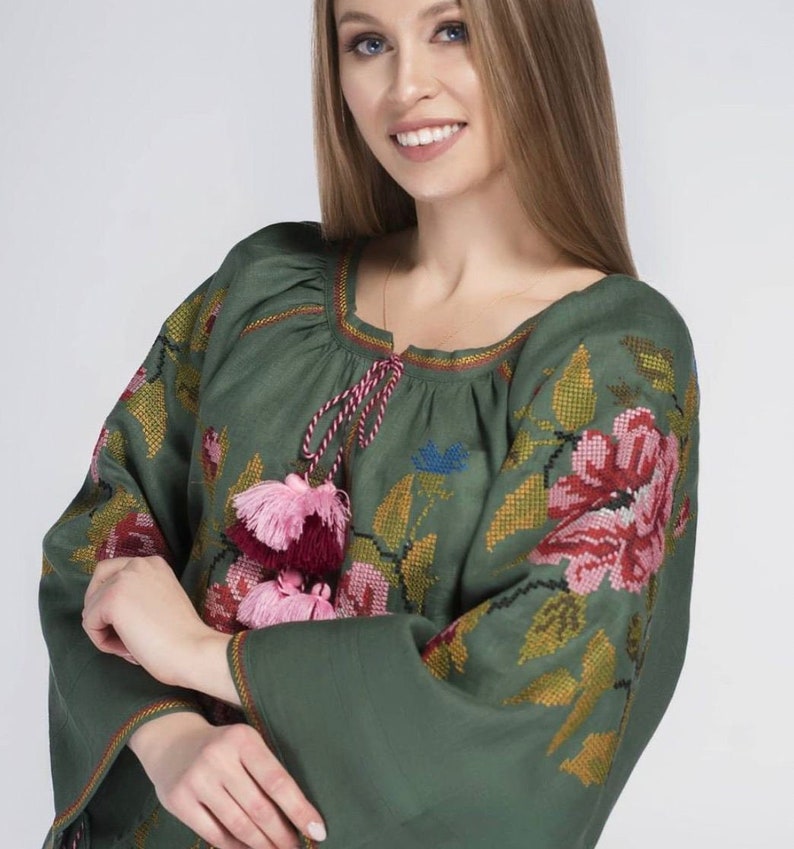 Puede incluir: Una blusa de lino verde con detalles florales bordados y borlas rosas y burdeos. La blusa tiene un escote redondo y mangas largas.