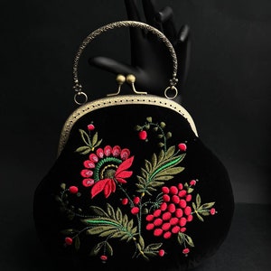 Pode incluir: Uma bolsa de m&atilde;o de veludo preto com um design de bordado floral em tons de vermelho e verde. A bolsa tem um fecho de lat&atilde;o e uma al&ccedil;a superior.