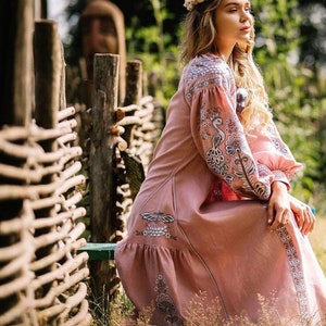 K&ouml;nnte beinhalten: Eine Frau in einem langen, rosafarbenen Leinenkleid mit Blumenstickerei. Sie sitzt auf einer Holzbank vor einem geflochtenen Zaun. Sie tr&auml;gt eine Blumenkrone im Haar.