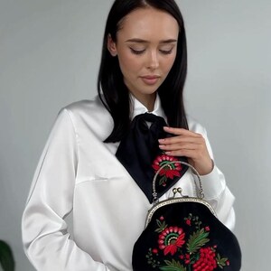Pode incluir: Uma mulher vestindo uma blusa branca com uma gravata borboleta preta segura uma embreagem de veludo preto com flores bordadas vermelhas e verdes.